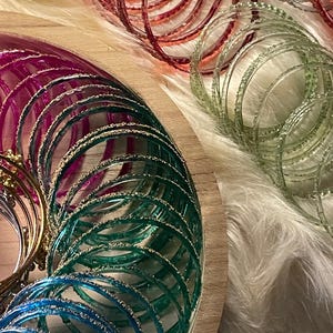 May include: A selection of colourful bangles in blue, green, pink, and red, some with glitter detailing. The bangles are displayed on a wooden surface and a fluffy white background.