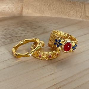 May include: A set of three gold-toned rings. One ring features a bamboo-like texture with small clear stones. Another is a textured band with tiny red stones. The third is a wide, woven-style band with blue flower accents and a central red stone.