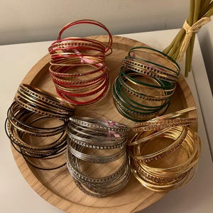 May include: A collection of thin metal bangles arranged in five distinct stacks. The sets feature red, dark green, black, silver, and gold tones, with many of the individual bracelets accented by small, sparkling embedded stones along their outer edges.