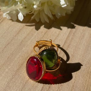 May include: Two gold rings, each with a large, oval gemstone. One ring has a bright red stone, the other a green one. The rings are on a wooden surface, with a white flower in the background. The gemstones are approximately 2 cm in length.