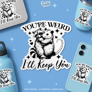 May include: A collection of stickers and accessories featuring a raccoon and opossum hugging, with the text "You're Weird, I'll Keep You." The stickers are black and white, with a blue background. Other items include a laptop, phone, and water bottles.