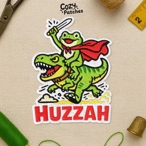 May include: Embroidered patch featuring a green frog riding a green dinosaur, both outlined in white. The frog, wearing a red cape and holding a sword, is above the word "Huzzah" in red. The patch has a cartoon style.