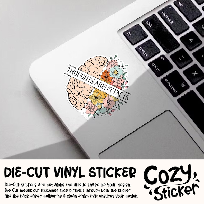 May include: A die-cut vinyl sticker featuring a stylized brain graphic with colorful flowers. The sticker has the text "THOUGHTS AREN'T FACTS" across the brain. The sticker is on a white laptop surface.