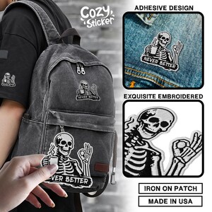 Puede incluir: Parches bordados en blanco y negro con un dise&ntilde;o de esqueleto sosteniendo una taza de caf&eacute; y el texto "NEVER BETTER." Los parches son para planchar y se muestran en una chaqueta vaquera y una mochila. La imagen tambi&eacute;n muestra las palabras "Cozy Sticker."