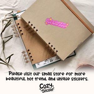 Puede incluir: Una pila de cuadernos con espiral con una pegatina rosa y morada que dice: "Do You Guys Ever Think About Dying?". Los cuadernos son marrones y beige con encuadernaci&oacute;n en espiral dorada.