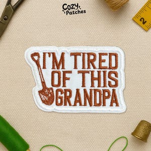 May include: Embroidered patch with the text "I'M TIRED OF THIS GRANDPA" in brown, featuring a shovel design. The patch has a white border and is likely for clothing or accessories. The patch is from Cozy Patches.