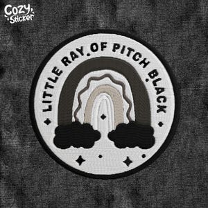 May include: Embroidered patch with a rainbow design in shades of black, gray, and beige, with the text "LITTLE RAY OF PITCH BLACK" in a circular pattern. The patch includes black clouds and star accents. The Cozy Sticker logo is in the upper left corner.