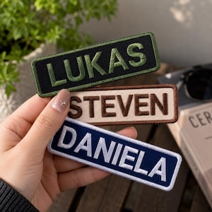 May include: Three rectangular embroidered name patches with stitched borders. The top patch is black with olive green text reading LUKAS. The middle patch is beige with brown text reading STEVEN. The bottom patch is navy blue with white text reading DANIELA.