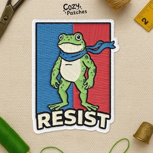 May include: Embroidered patch featuring a green frog wearing a blue scarf, set against a red and blue background. The word "RESIST" is embroidered in white below the frog. The patch is from Cozy Patches.