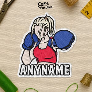 May include: Embroidered patch featuring a female boxer in a red top and blue boxing gloves. The patch has the text "ANYNAME" in bold white letters. The patch is outlined in white and has the text "Cozy Patches" in the top right corner.