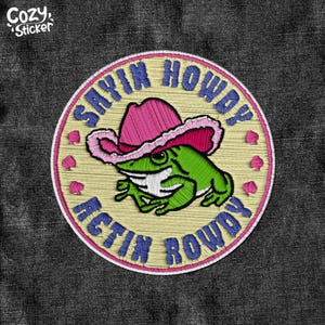 Peut inclure: Un patch brodé jaune et rose avec une grenouille verte portant un chapeau de cowboy rose. Le texte sur le patch est "Sayin Howdy Actin Rowdy".
