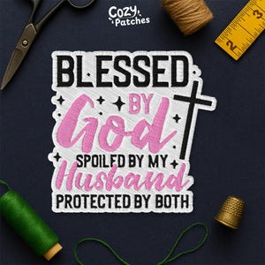 May include: Embroidered patch with the text "BLESSED BY GOD, SPOILED BY MY HUSBAND, PROTECTED BY BOTH" in black and pink. The patch has a white border and a cross design. The patch is surrounded by sewing tools and accessories.