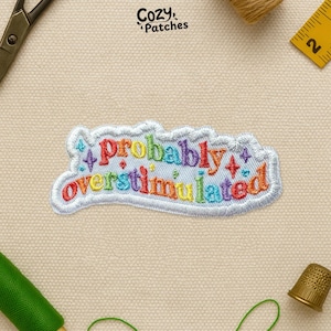May include: Embroidered patch with the phrase "probably overstimulated" in rainbow colors. The patch has a white border and is decorated with small star-like shapes. The brand name "Cozy Patches" is in the top right corner.