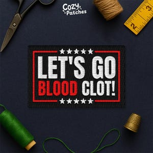 May include: Black rectangular patch with the text "LET'S GO BLOOD CLOT!" in white and red, framed by a red border and stars. The patch is surrounded by sewing tools and supplies, including scissors, thread, and a thimble.
