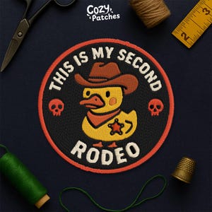 May include: Embroidered patch featuring a yellow duck wearing a cowboy hat and bandana, with a sheriff's star. The patch reads "THIS IS MY SECOND RODEO" in white lettering on a black background, with a red border and two skull designs.