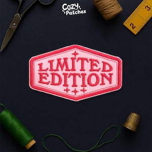 May include: A pink and red embroidered patch with the words "LIMITED EDITION" in a bold font. The patch has a decorative border and small cross-like symbols. The patch is surrounded by sewing tools and supplies, including scissors, thread, and a thimble.