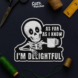 Peut inclure: Écusson brodé noir et blanc représentant un squelette tenant une tasse, avec le texte "AS FAR AS I KNOW I'M DELIGHTFUL". L'image comprend des outils de couture tels que des ciseaux, du fil et un dé à coudre.