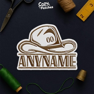 May include: Embroidered patch featuring a cowboy hat design with the text "ANYNAME" below. The hat is brown and white, with the number "00" on the front. The patch is surrounded by sewing tools, including scissors, a measuring tape, and thread.