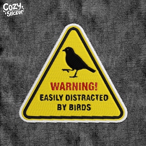 May include: Embroidered patch with a yellow triangle warning sign featuring a black bird silhouette. The text "WARNING! EASILY DISTRACTED BY BIRDS" is in red and black. The patch has a white border and the text "Cozy Sticker" in the upper left corner.