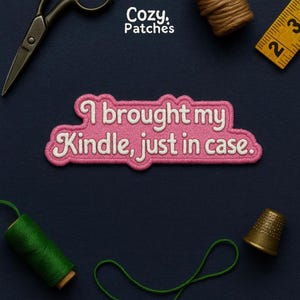 May include: A pink embroidered patch with the text "I brought my Kindle, just in case." The patch has a white border and is surrounded by sewing tools, including scissors, thread, and a measuring tape. The brand name "Cozy Patches" is at the top.