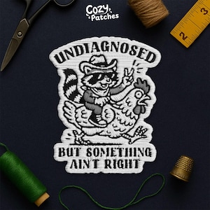 Peut inclure: Écusson brodé avec un raton laveur portant des lunettes de soleil et un chapeau de cowboy, chevauchant une poule. Le texte dit "UNDIAGNOSED" et "BUT SOMETHING AIN'T RIGHT". Le patch est blanc avec des détails noirs.