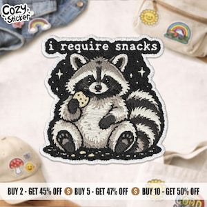 May include: An embroidered patch featuring a gray and white raccoon sitting on a black background, holding a small cookie. The text "i require snacks" is stitched in white above the raccoon, surrounded by small white star-shaped accents.