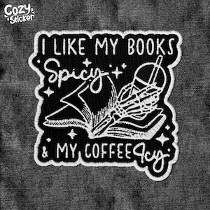 May include: Black and white embroidered patch with the text "I like my books spicy & my coffee cozy". The patch features a stylized image of a book with a skeleton hand holding a quill pen.