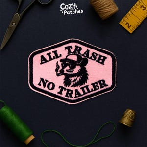 May include: A pink and black embroidered patch featuring the text "ALL TRASH NO TRAILER" and a raccoon wearing sunglasses and a hat, smoking. Sewing tools and thread surround the patch.