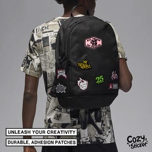 May include: Black backpack adorned with colorful patches, including text and graphic designs. The patches feature phrases like "ALL TRASH NO TRAILER" and "WORLD CHAMPIONS". The backpack has a side pocket and a top handle. The text "UNLEASH YOUR CREATIVITY" and "DURABLE, ADHESION PATCHES" are also visible.