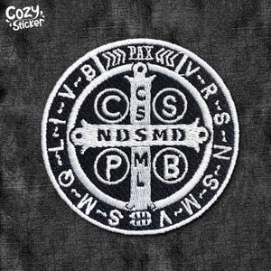 Peut inclure: Un patch brodé noir et blanc d'une médaille de Saint Benoît. La médaille représente une croix avec les lettres "C.S.P.B." et "N.D.S.M.D." autour d'elle. Les mots "V.R.S.N." et "PAX" sont autour de l'extérieur de la médaille.