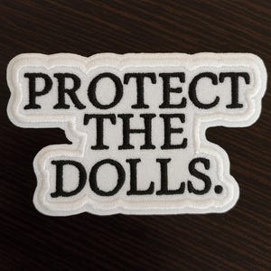 Peut inclure: Écusson brodé blanc avec le texte noir "PROTECT THE DOLLS". Le patch a une forme irrégulière et une bordure blanche. Le fond est une surface marron foncé. Le texte est en gras.