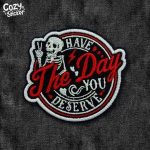 May include: Embroidered patch with a skeleton giving a peace sign, surrounded by a red and white circular border. The patch reads "Have The Day You Deserve" in red script and white lettering on a black background.