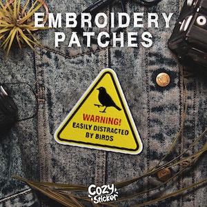 Pode incluir: Patch de bordado triangular amarelo com uma silhueta de p&aacute;ssaro preto e o texto "WARNING! EASILY DISTRACTED BY BIRDS." O patch est&aacute; em uma jaqueta jeans. As palavras "EMBROIDERY PATCHES" est&atilde;o no topo.