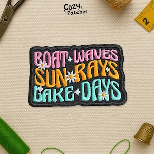 May include: Embroidered patch with the words "Boat Waves Sun Rays Lake Days" in pink, orange, and teal, with daisy accents. The patch has a black border and the brand name "Cozy Patches" is in the top left corner.