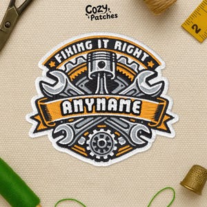 May include: Embroidered patch with the text "FIXING IT RIGHT" and "ANYNAME". The design features crossed wrenches, a piston, and a gear, with orange and gray accents. The patch is on a beige fabric background.