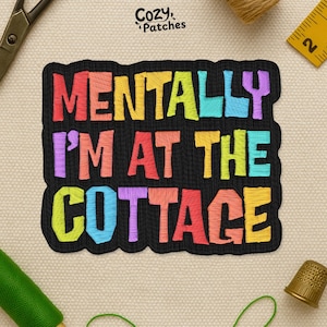 May include: Embroidered patch with the text "MENTALLY I'M AT THE COTTAGE" in colorful letters on a black background. The patch is surrounded by sewing tools, including scissors, a thimble, and thread.