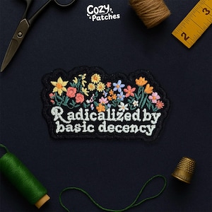 Peut inclure: Écusson brodé noir avec des fleurs colorées et le texte "Radicalized by basic decency". L'image comprend des outils de couture, tels que des ciseaux, un dé à coudre et un mètre ruban, sur un fond bleu foncé. La marque "Cozy Patches" est visible.