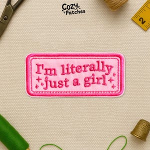 May include: A pink embroidered patch with the text "I'm literally just a girl" in a rectangular shape. The patch has a pink border and is decorated with small star-like symbols. The brand name "Cozy Patches" is in the top left corner.