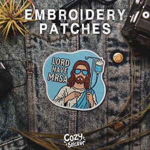 Puede incluir: Parche bordado con el texto "LORD HAVE MRSA" con una ilustraci&oacute;n de Jes&uacute;s con gafas de sol y sosteniendo un goteo intravenoso. El parche es azul claro con detalles en blanco y beige. El logotipo de Cozy Sticker est&aacute; en la parte inferior.