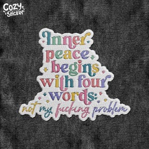 May include: A white embroidered patch with the text "Inner peace begins with four words: not my fucking problem" in various colors. The patch has a whimsical, irregular shape and the words are in a playful font. The patch is on a dark gray denim background.