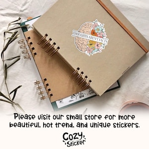May include: A sticker with a brain design, half in tan and half with colorful flowers, and the text "THOUGHTS AREN'T FACTS". The sticker is on a tan notebook with a spiral binding. The image also includes the text "Please visit our small store for more beautiful, hot trend, and unique stickers."