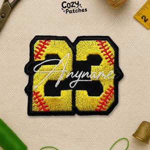 May include: Embroidered patch with the text "23" in yellow, black, and red baseball stitching. The word "Anyname" is embroidered in white script across the numbers. The patch is from Cozy Patches.