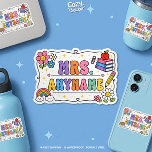 May include: A colorful sticker with the text "MRS. ANYNAME" in a retro font, surrounded by flowers, a rainbow, an apple, books, and a pencil. The sticker is displayed on various light blue items, including a laptop, a phone, and a water bottle.