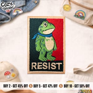 May include: Embroidered patch with a green frog wearing a blue scarf, set against a red and black backdrop. The word "RESIST" is stitched below the frog. The patch has a beige border, suitable for clothing or accessories.