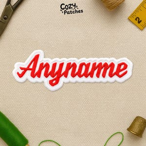 May include: Embroidered patch with the word "Anyname" in red script lettering, outlined in white. The patch is rectangular and has a stitched border. The brand name "Cozy Patches" is in the upper right corner.