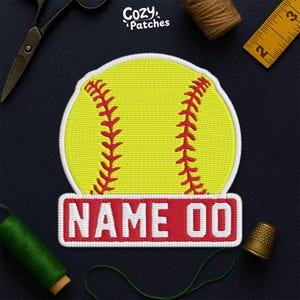 May include: Embroidered patch featuring a yellow softball with red stitching and a white border. Below the softball is a red rectangle with the text "NAME 00" in white. The patch is surrounded by sewing tools.