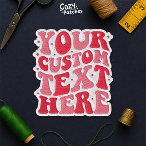 May include: Embroidered patch with the text "YOUR CUSTOM TEXT HERE" in shades of red and pink. The patch is surrounded by a white border. The image also includes sewing tools like scissors, a thimble, and measuring tape.
