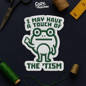 Peut inclure: Écusson brodé avec un motif de grenouille verte portant des lunettes et le texte "I MAY HAVE A TOUCH OF THE 'TISM". Le patch est blanc avec des coutures vertes. Le logo Cozy Patches est en haut à gauche.