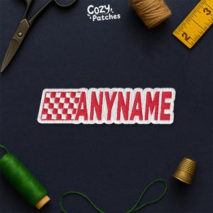 May include: Embroidered patch with the text "ANYNAME" in red, outlined in white, with a checkered flag design on the left. The patch is rectangular and likely for clothing or accessories. The brand name "Cozy Patches" is in the top right.