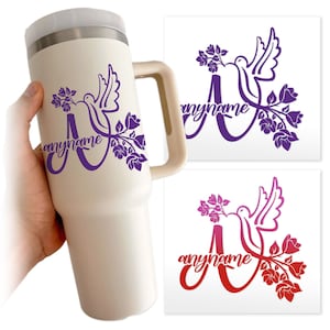 May include: A cream-colored insulated tumbler with a handle, featuring a purple dove and floral design with the word "anyname". Two additional designs in purple and red are shown on white backgrounds.